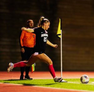 Female soccer player in a black jersey (number 25) kicks the ball from the corner near a yellow flag, with a referee in orange in the background.